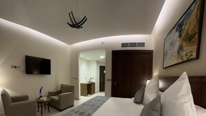 Quarto de hotel moderno com uma cama grande, duas poltronas, uma TV de tela plana e um quadro na parede.