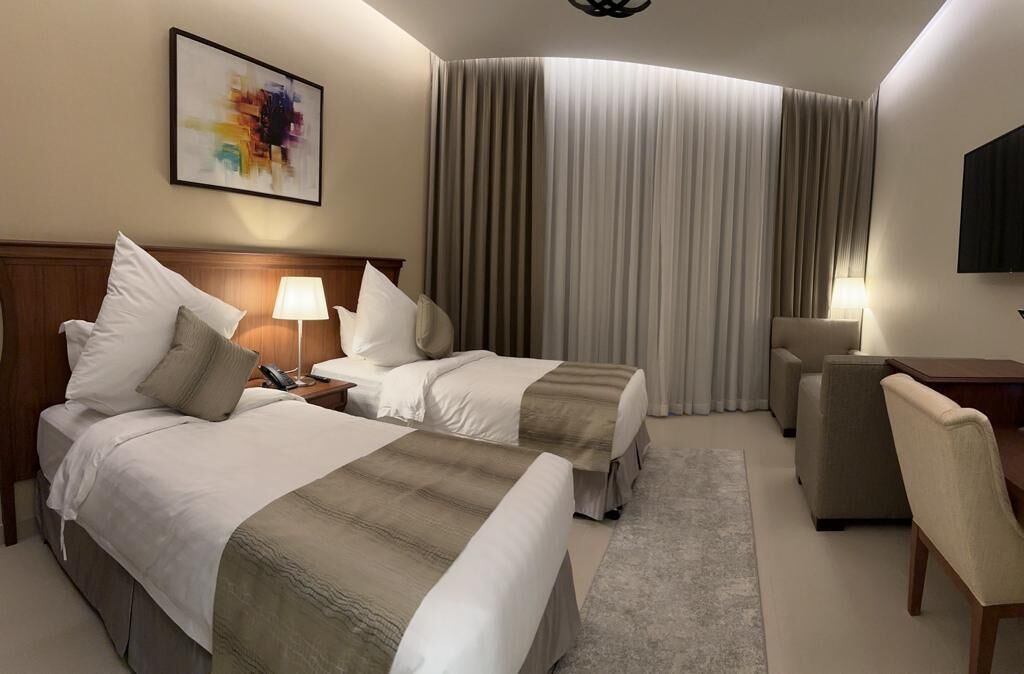 A hotel room with two beds, a desk, a chair, a TV, and a large window with curtains.