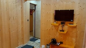 Habitación con paneles de madera, televisor montado en la pared, cafetera y disposición de asientos que incluye un sofá y dos otomanas.