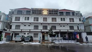 Un hotel de varios pisos con un letrero, un acceso cubierto y un estacionamiento con coches.