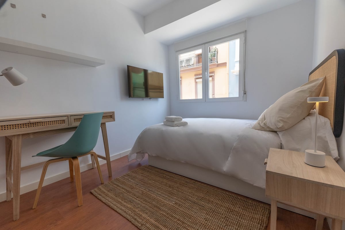 Ein Schlafzimmer mit Bett, Schreibtisch, Stuhl, Fernseher und Fenster.