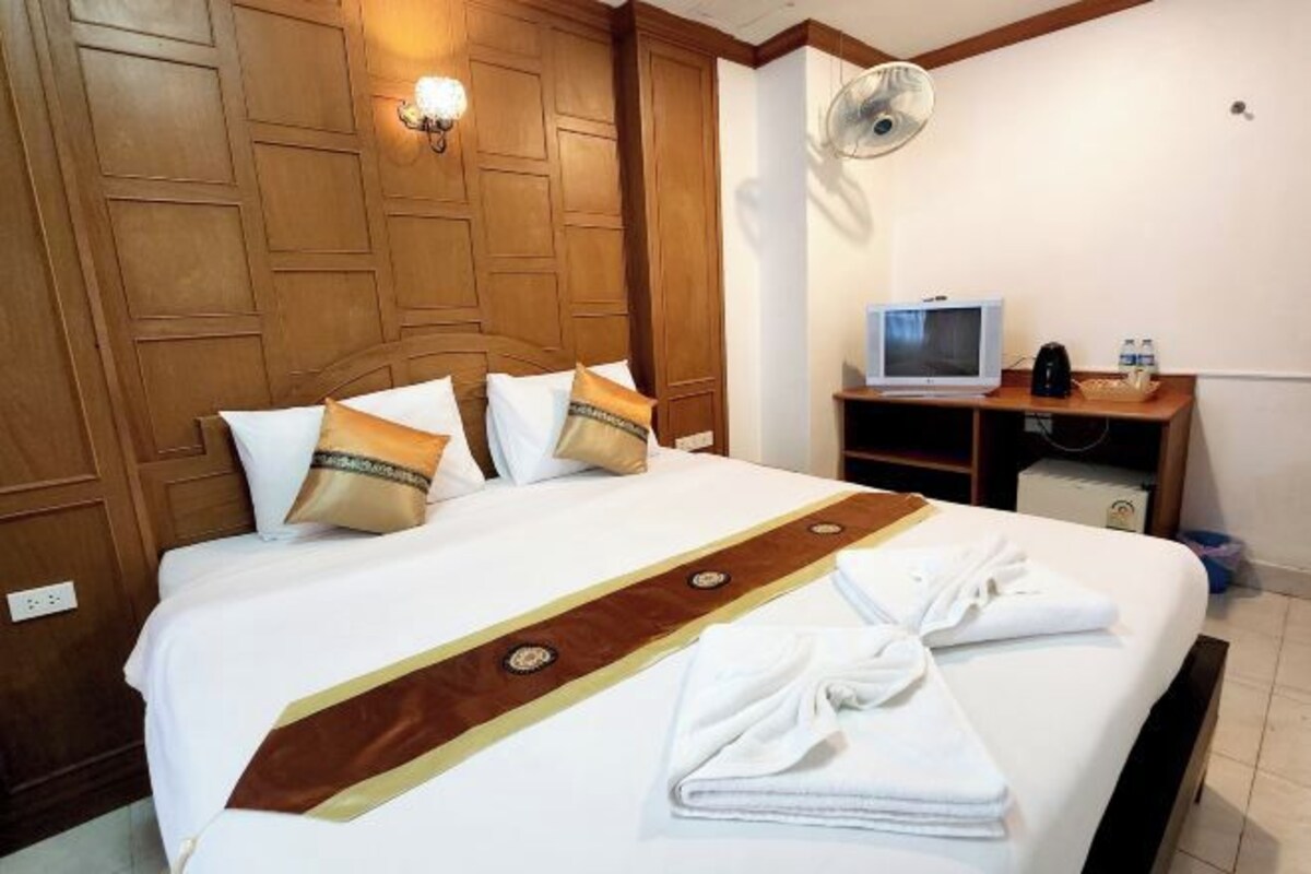 A hotel room with a wooden headboard, a bed with white bedding and a brown decorative strip, a television on a wooden stand, and a fan on the wall.