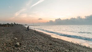 Una playa de guijarros con una persona pescando, mar tranquilo y cielo al atardecer.