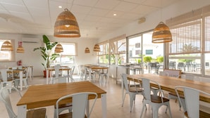 Un café amplio con mesas de madera y sillas blancas, ventanas grandes y lámparas de mimbre colgantes.