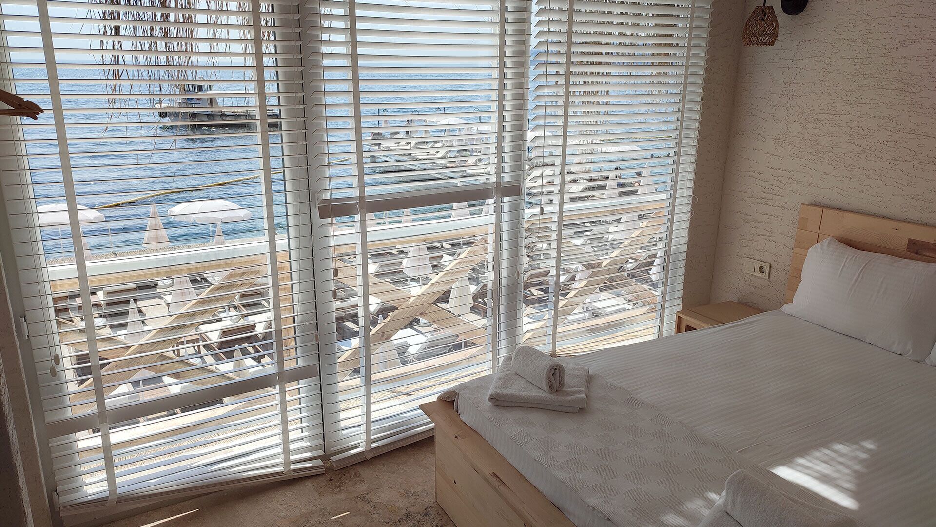 A hotel room with a large window overlooking a beach and boats. The room has a bed, a wooden headboard, and a small bedside table.