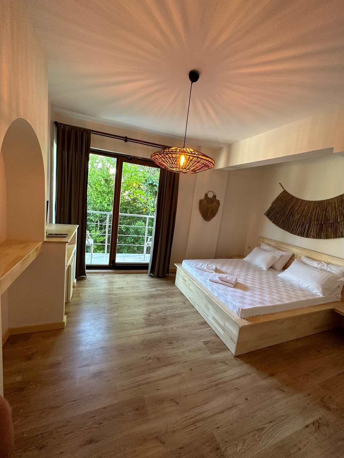 Un dormitorio con una cama grande, una lámpara colgante, vista a zonas verdes por la ventana y un balcón.