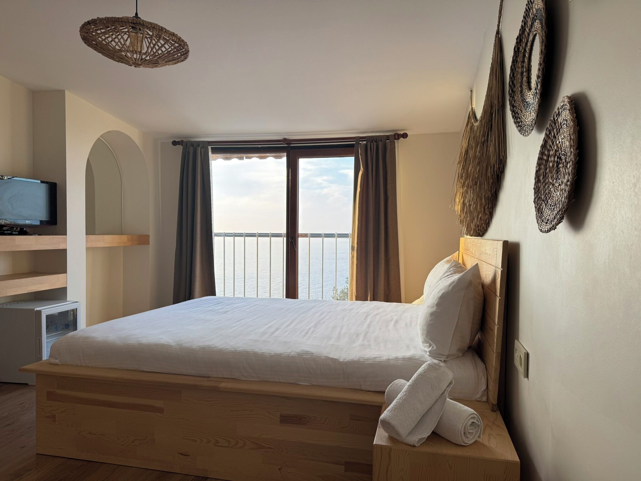 Ein Schlafzimmer mit einem hölzernen Bett, einem Fernseher auf einem Regal, einem Balkon mit Vorhängen und Blick auf das Meer.
