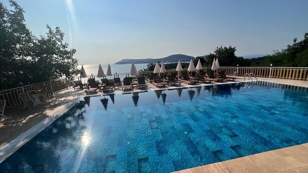 A clear blue infinity pool with lounge chairs and umbrellas, surrounded by a wooden deck and overlooking a body of water with distant landforms. - The Roof ADA (Adalar)