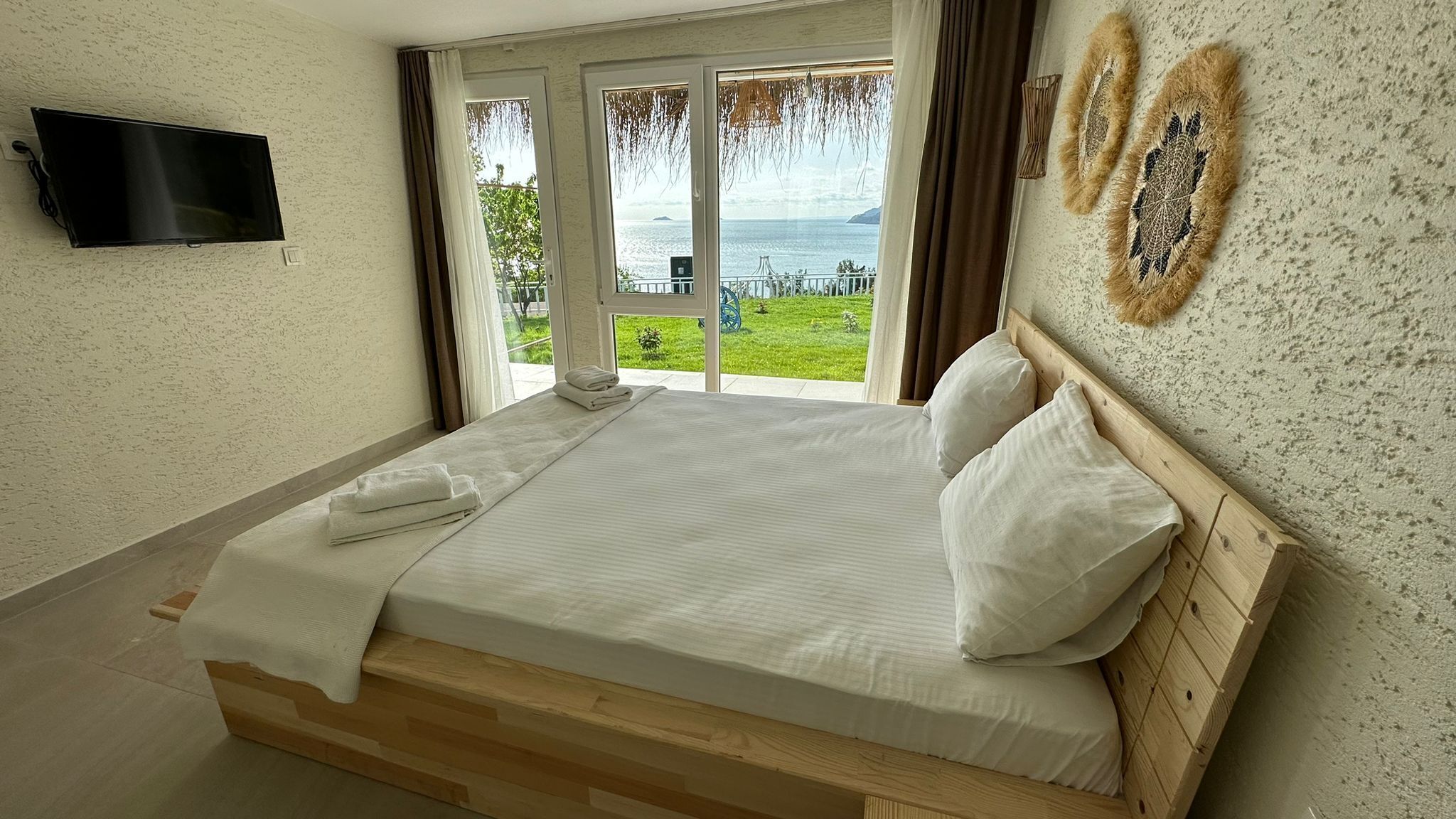 Een slaapkamer met een bed, een aan de muur bevestigde televisie en uitzicht op zee door grote ramen.