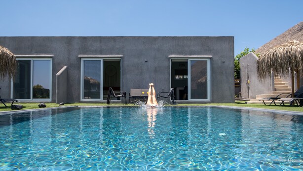 A swimming pool with clear blue water, surrounded by a modern building with large windows and a thatched roof structure. - Sagma Villas (Santorini)
