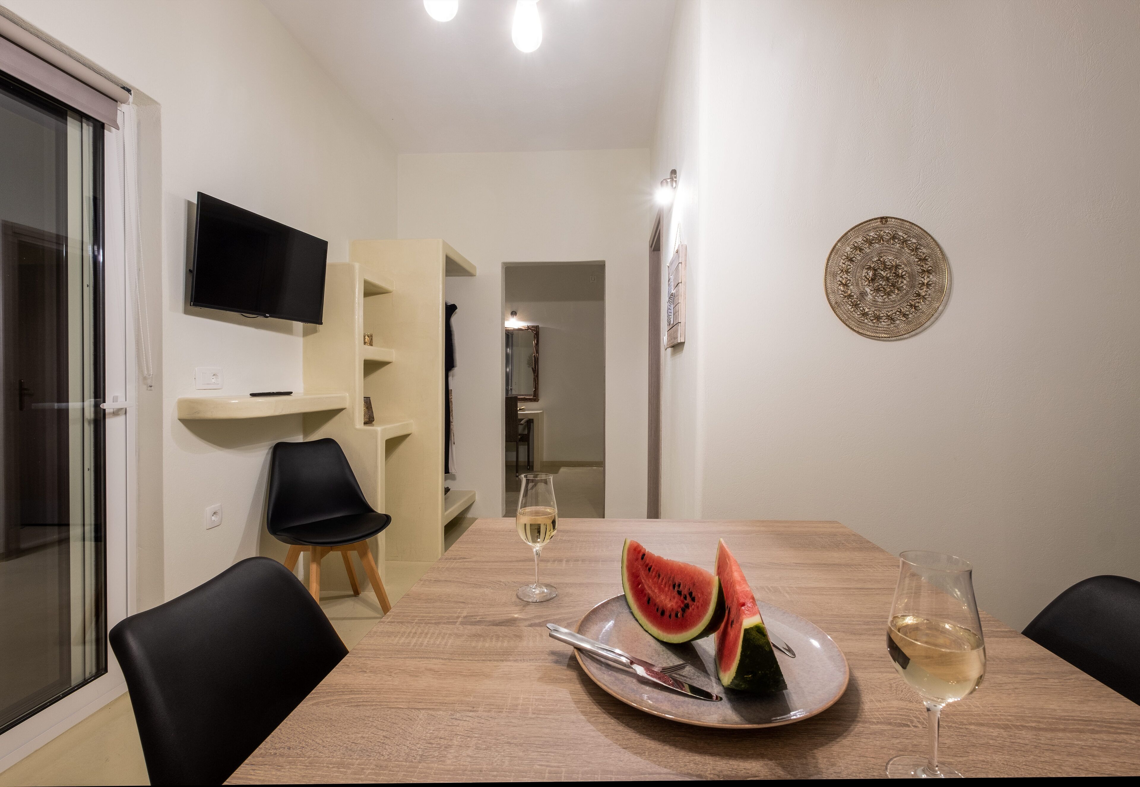 A dining area with a wooden table, two black chairs, a plate with watermelon slices, a wine glass, and a TV mounted on the wall.