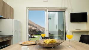A modern kitchen with a dining area, a large window with a mountain view, and a flat-screen TV mounted on the wall. - Sagma Villas (Santorini)