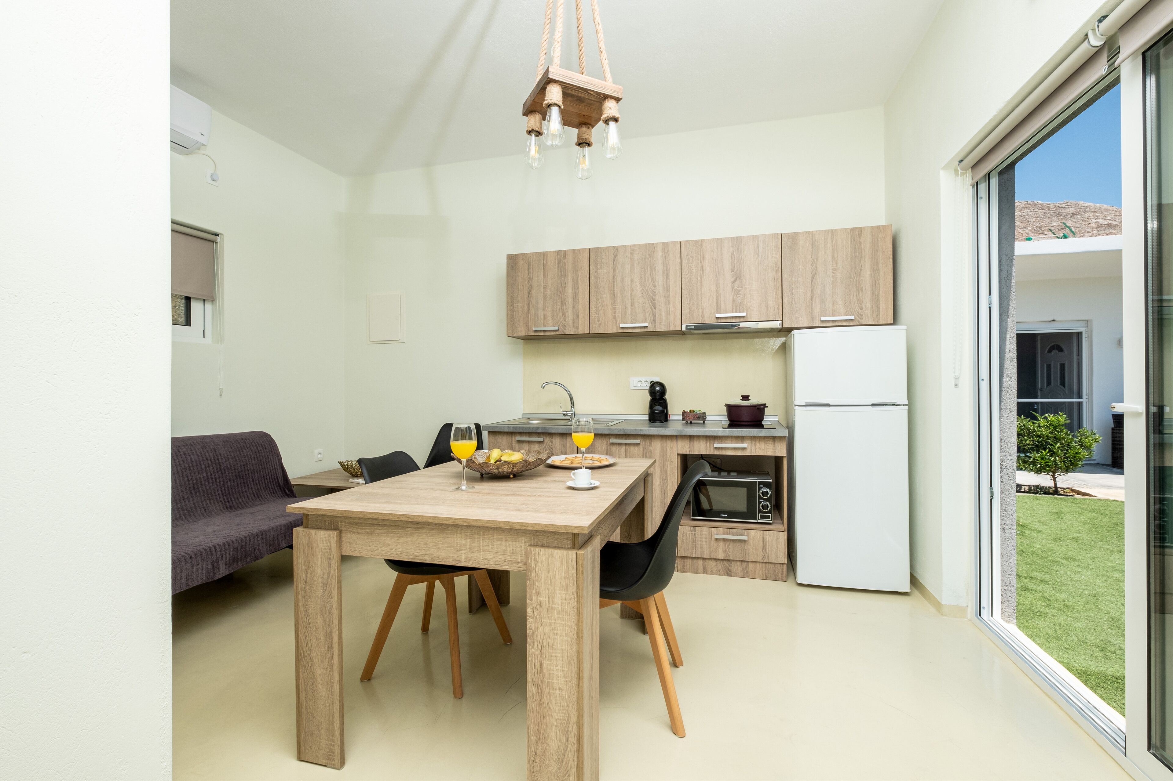 A modern kitchen with wooden cabinets, a dining table with chairs, a sofa, and a view of a green lawn through a sliding glass door.