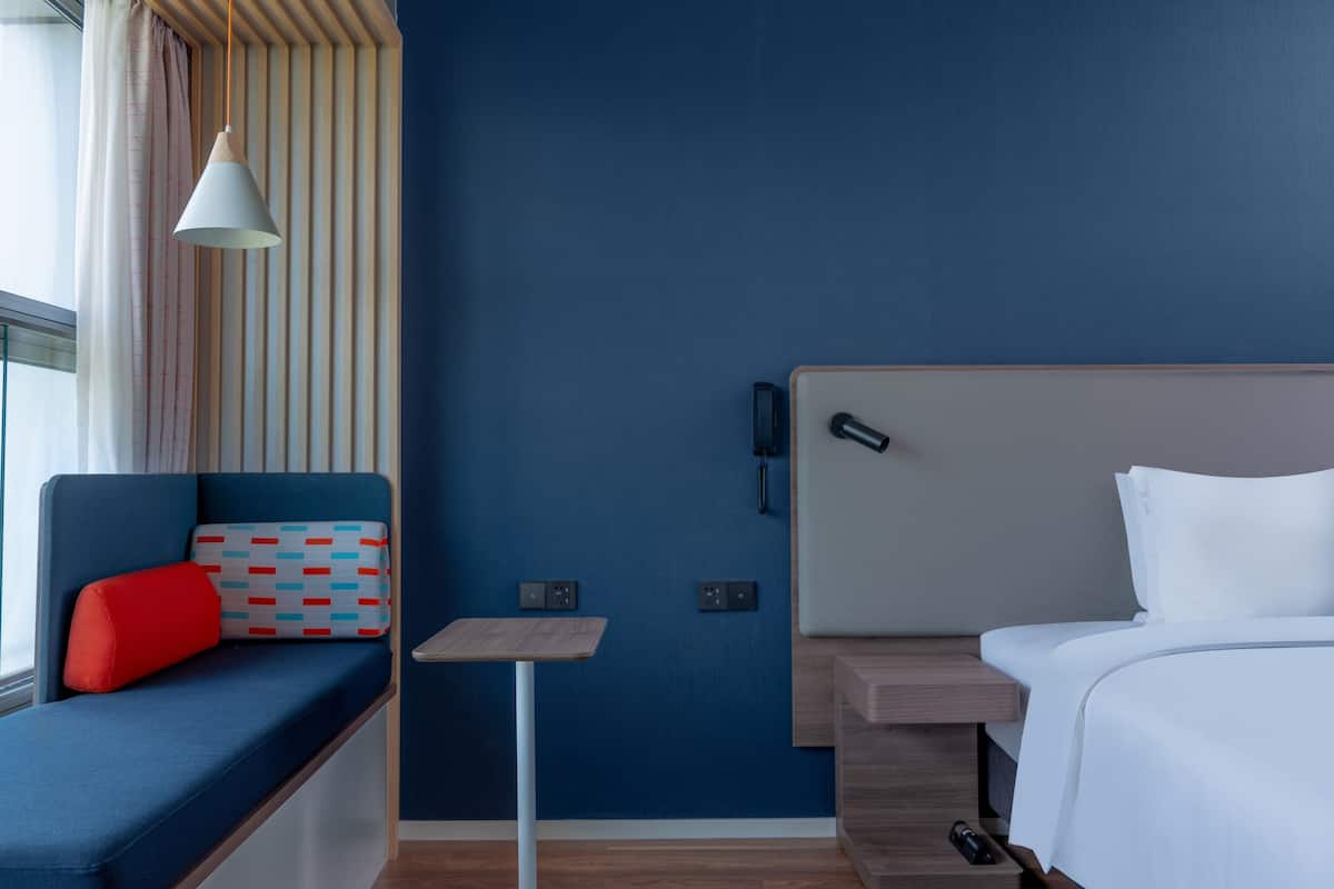 Ett modernt hotellrum med en blå accentvägg, en säng med vita sängkläder, ett litet bord och en sittgrupp med en röd kudde.