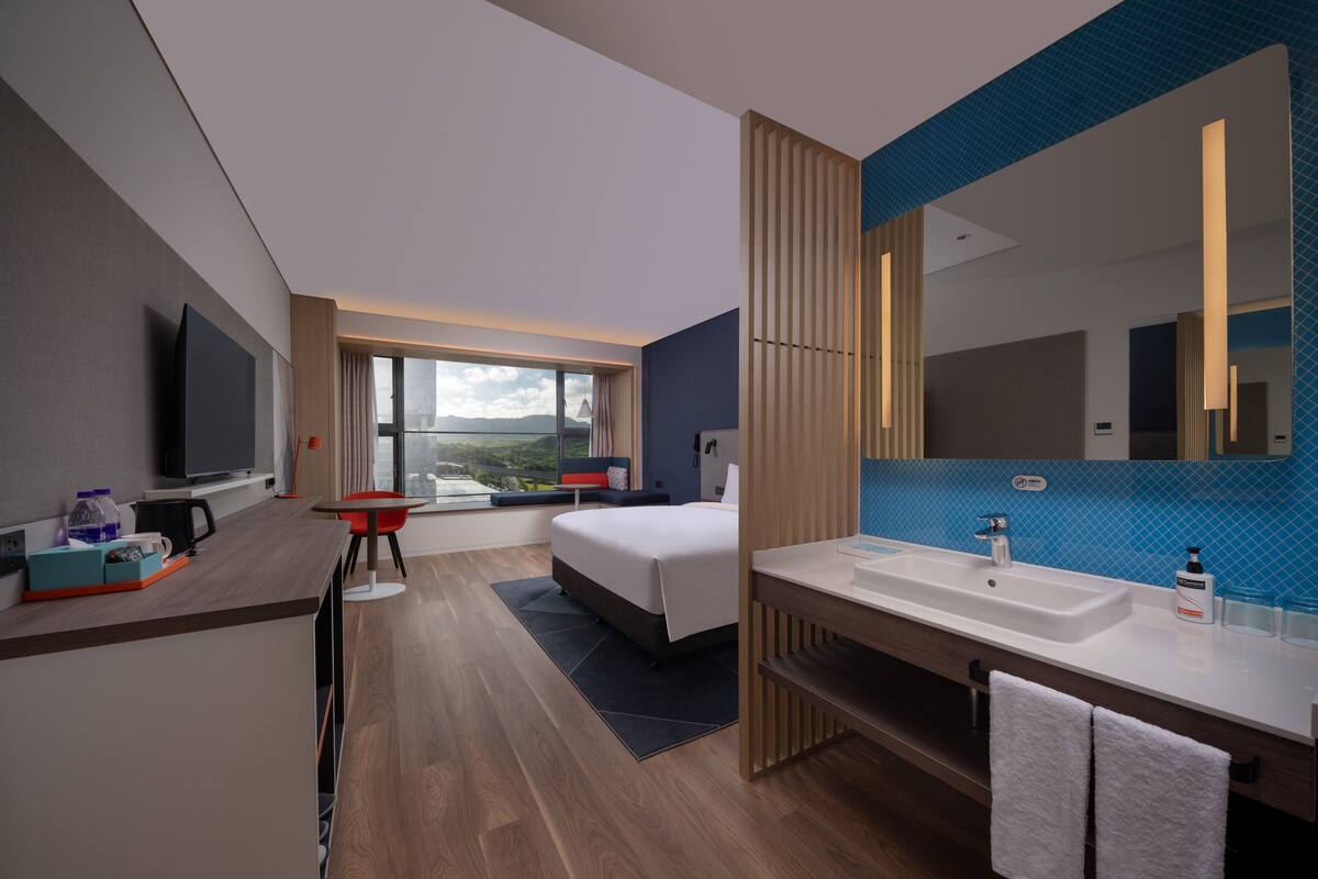 Ett modernt hotellrum med en stor säng, ett skrivbord med en stol, en TV, ett stort fönster med utsikt, ett badrum med handfat och spegel samt en blå kakelbeklädd vägg.