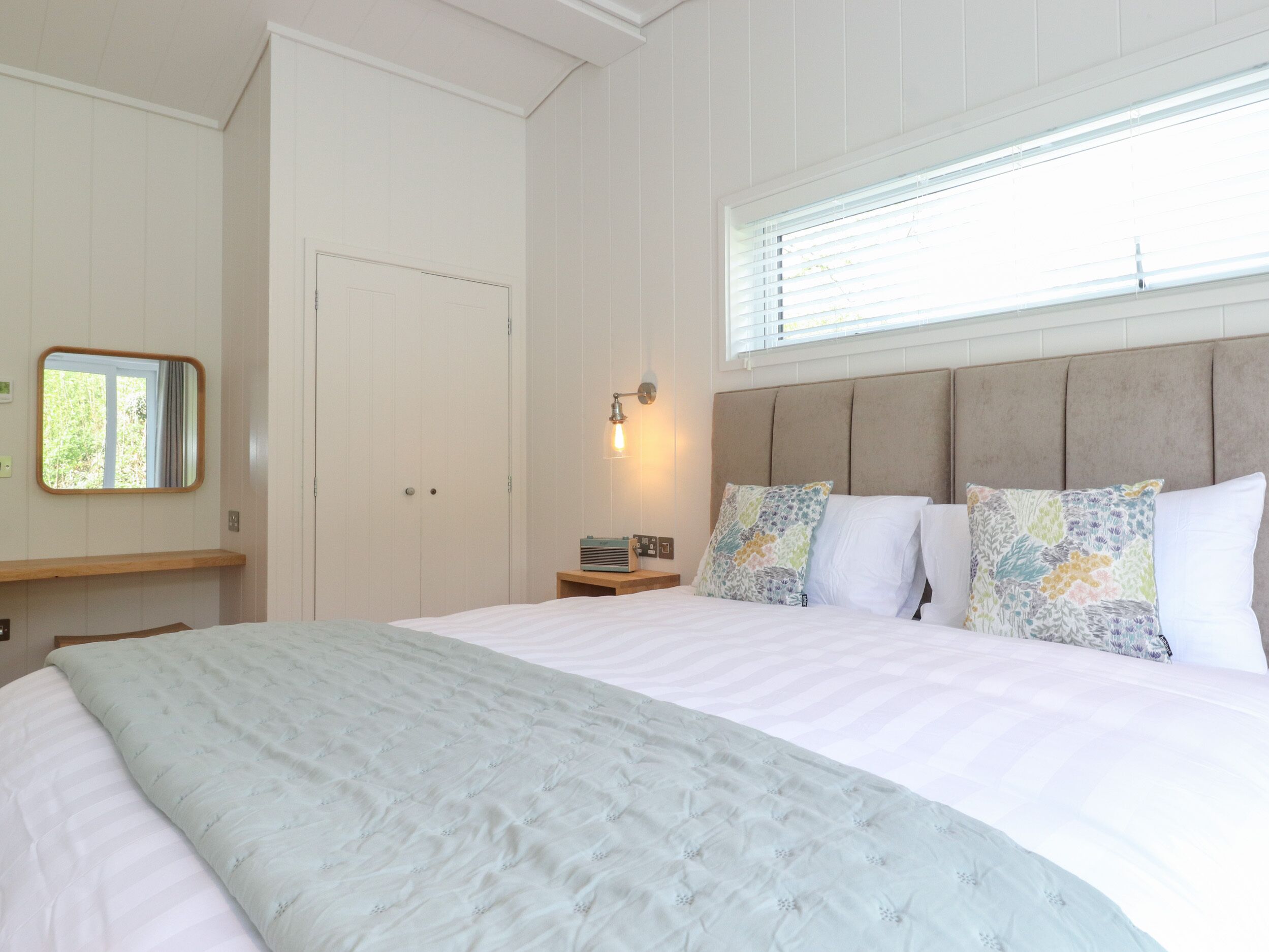 A bedroom with a large bed, a bedside table, a nightstand, a mirror, and a window with blinds.