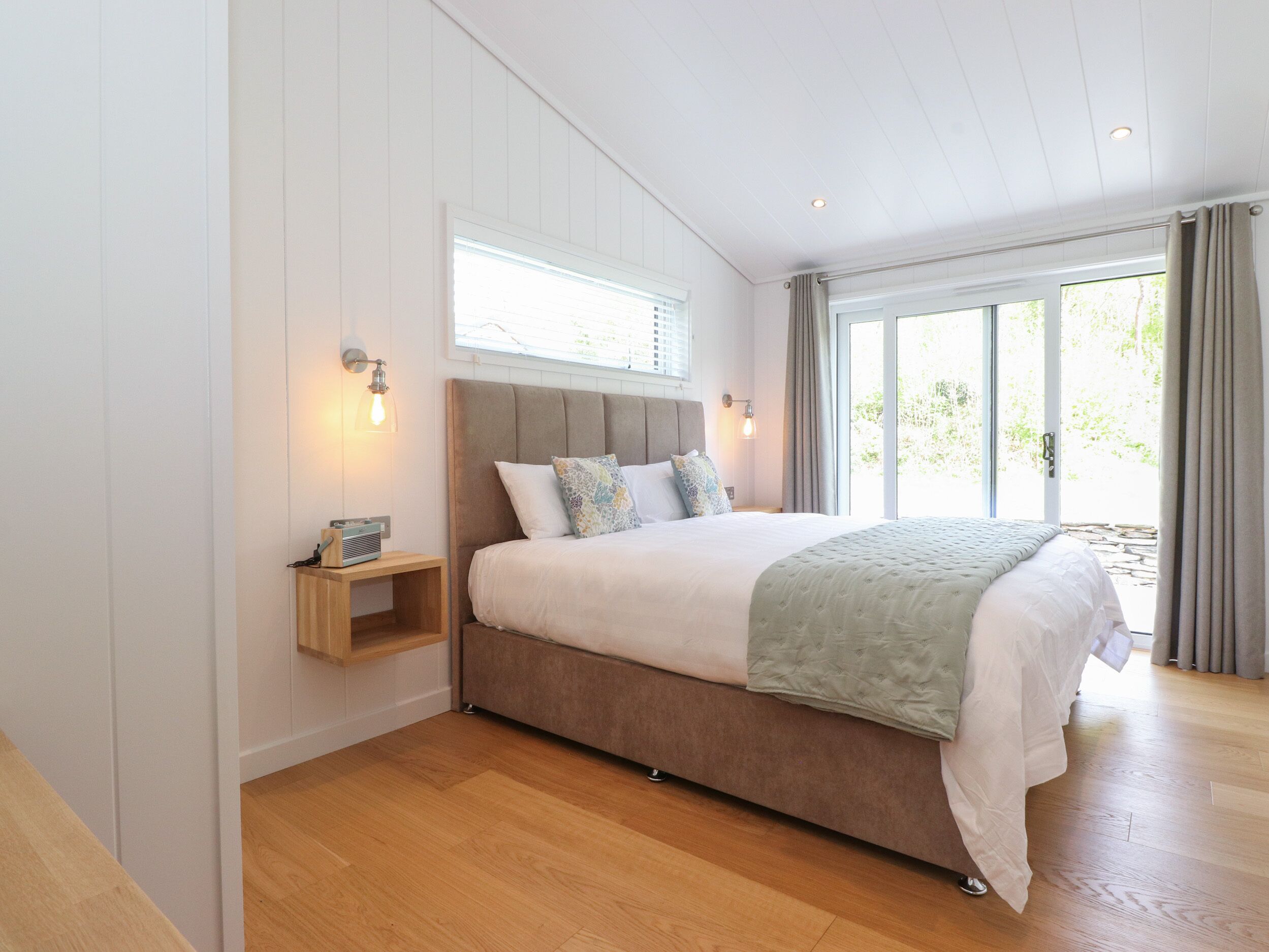 A bedroom with a large bed, wooden nightstands, and a view of greenery through the window.