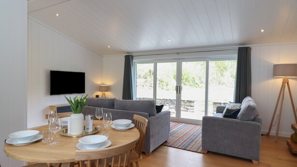 A modern living room with a wooden dining table, grey sofas, and a television mounted on the wall. - Hawthorn Croft (Ulverston)
