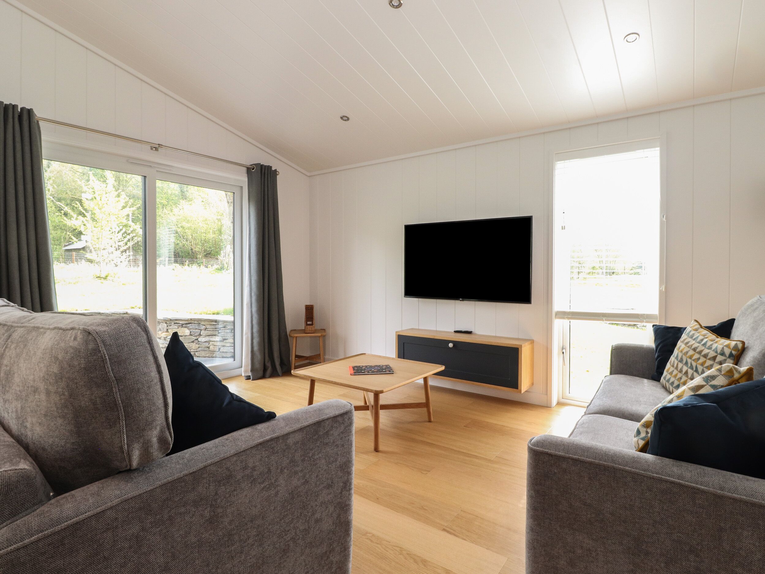 Een moderne woonkamer met een flatscreen televisie, twee grijze banken, een houten salontafel en een stenen muur.