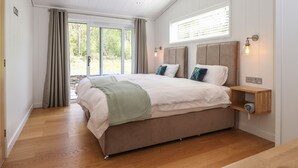 Un dormitorio con una cama grande, piso de madera, una ventana con cortinas y una mesita de noche con una lámpara.