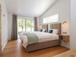 Un dormitorio con una cama grande, piso de madera, una ventana con cortinas y una mesita de noche con una lámpara.