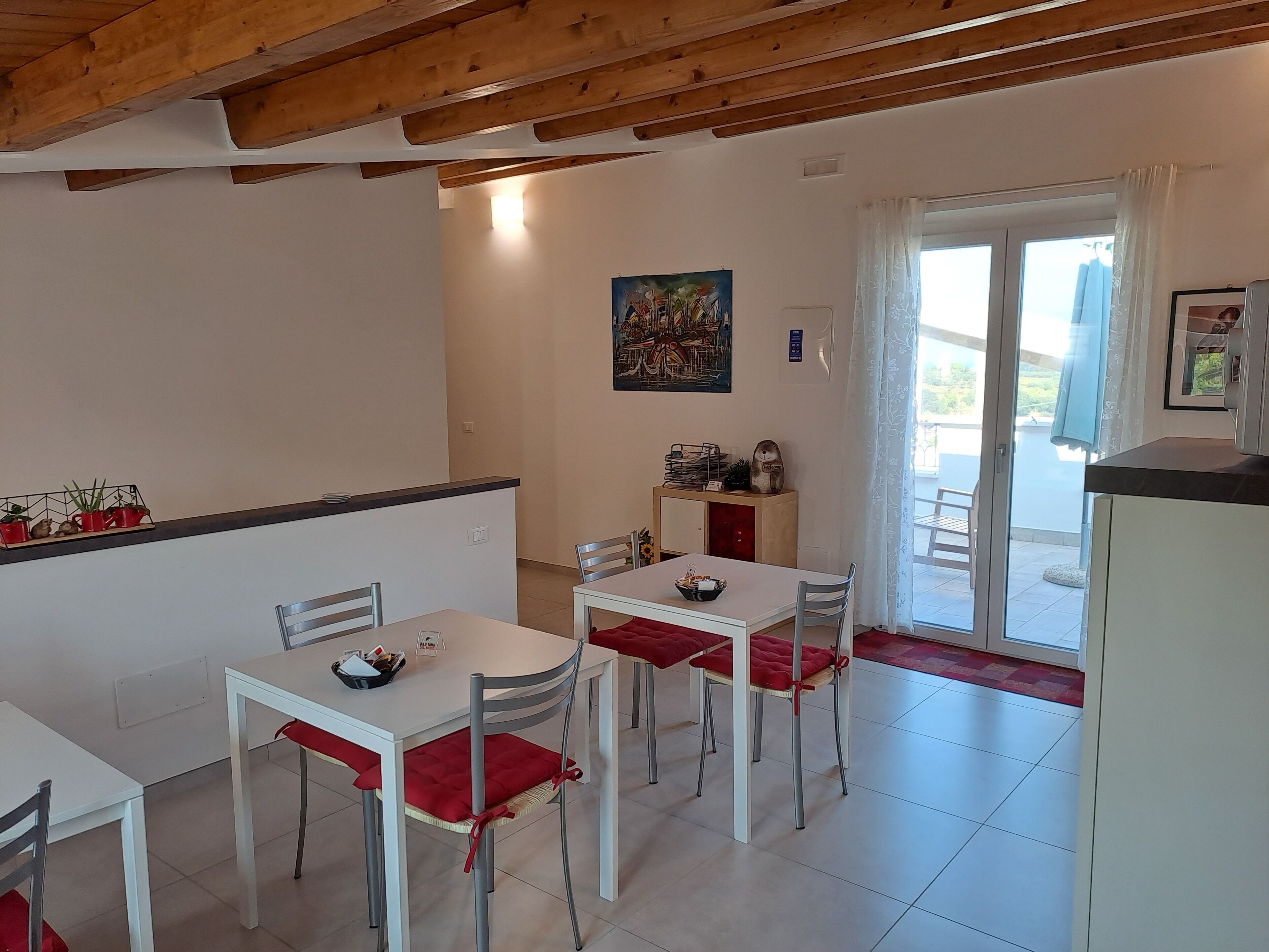 Un comedor con mesas y sillas blancas, una encimera de cocina y vistas al exterior a través de un ventanal.