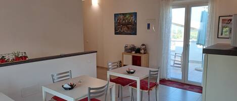 Un comedor con mesas y sillas blancas, una encimera de cocina y vistas al exterior a través de un ventanal.