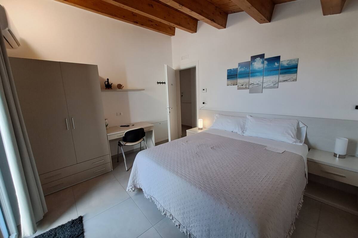 A bedroom with a bed, a desk, a wardrobe, and a wall with a painting.