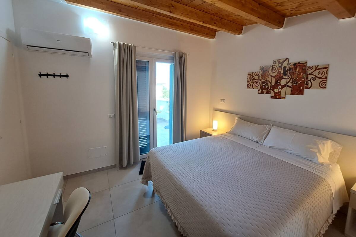 A bedroom with a bed, a desk, and a chair. There is a window with curtains, a ceiling with wooden beams, and a wall with a painting.