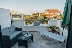 A balcony with a sofa, a table, and a sun lounger. There is a view of a building and trees in the background. - B&B TANA DEL RICCIO (Ortona)