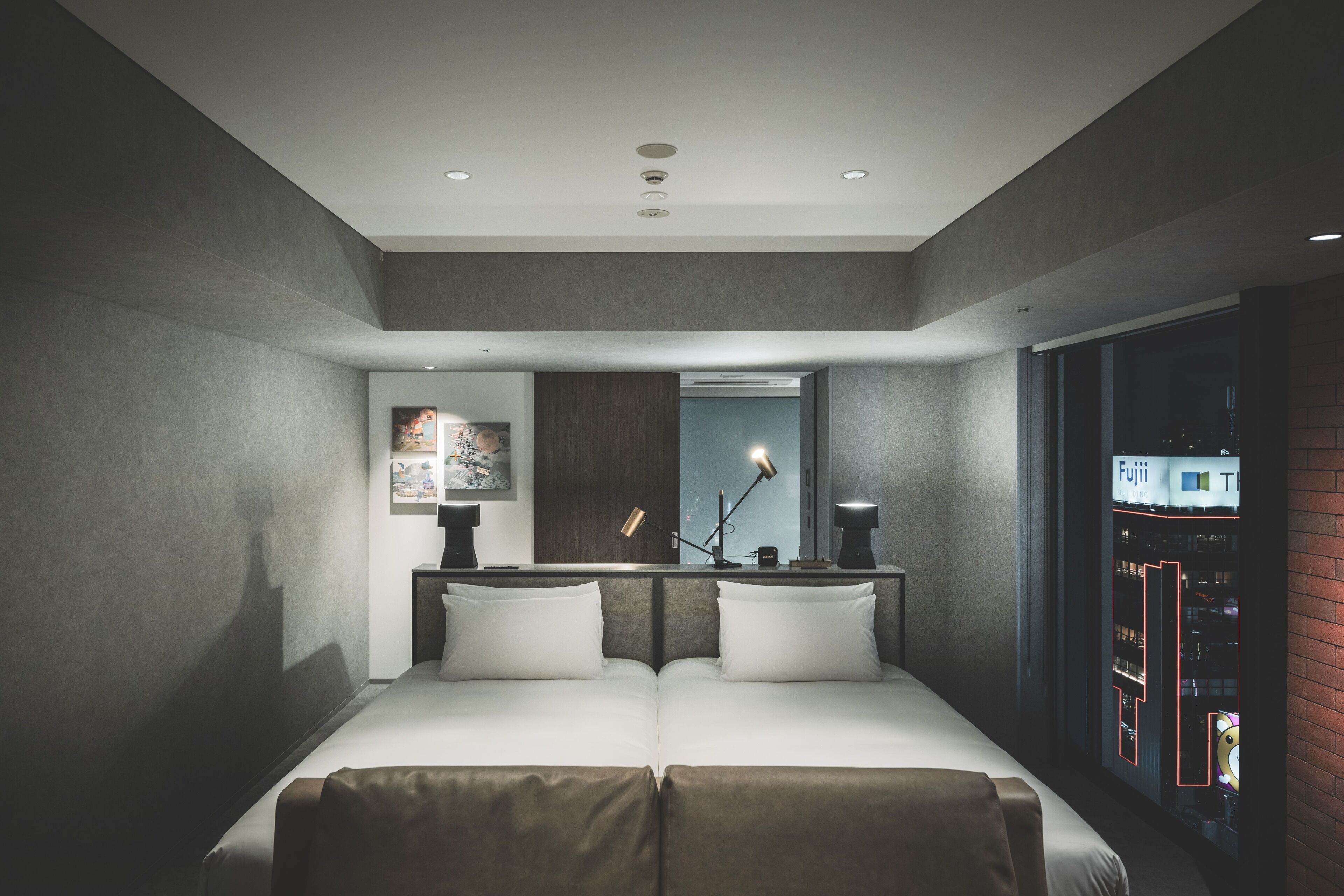 A modern hotel room with a large bed, a nightstand, a mirror, and a view of the city through a window.