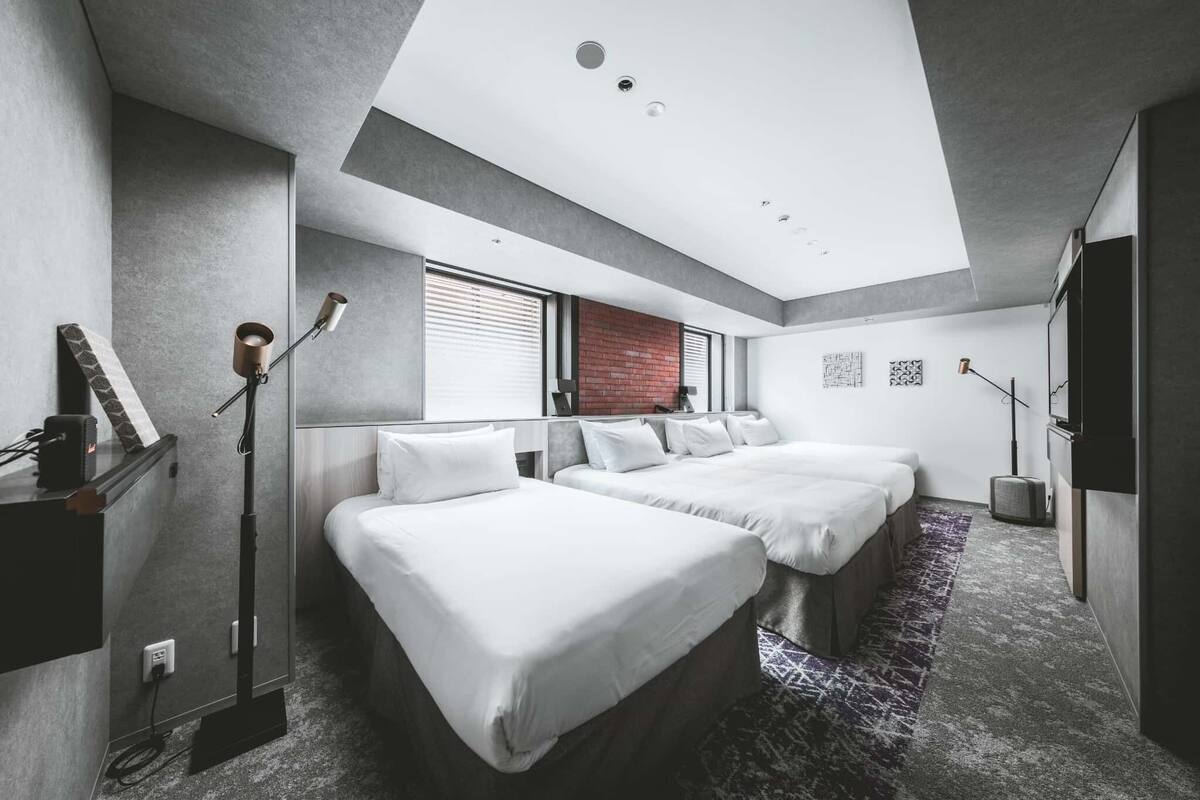 A modern hotel room with three beds, a skylight, a brick accent wall, and a large window.
