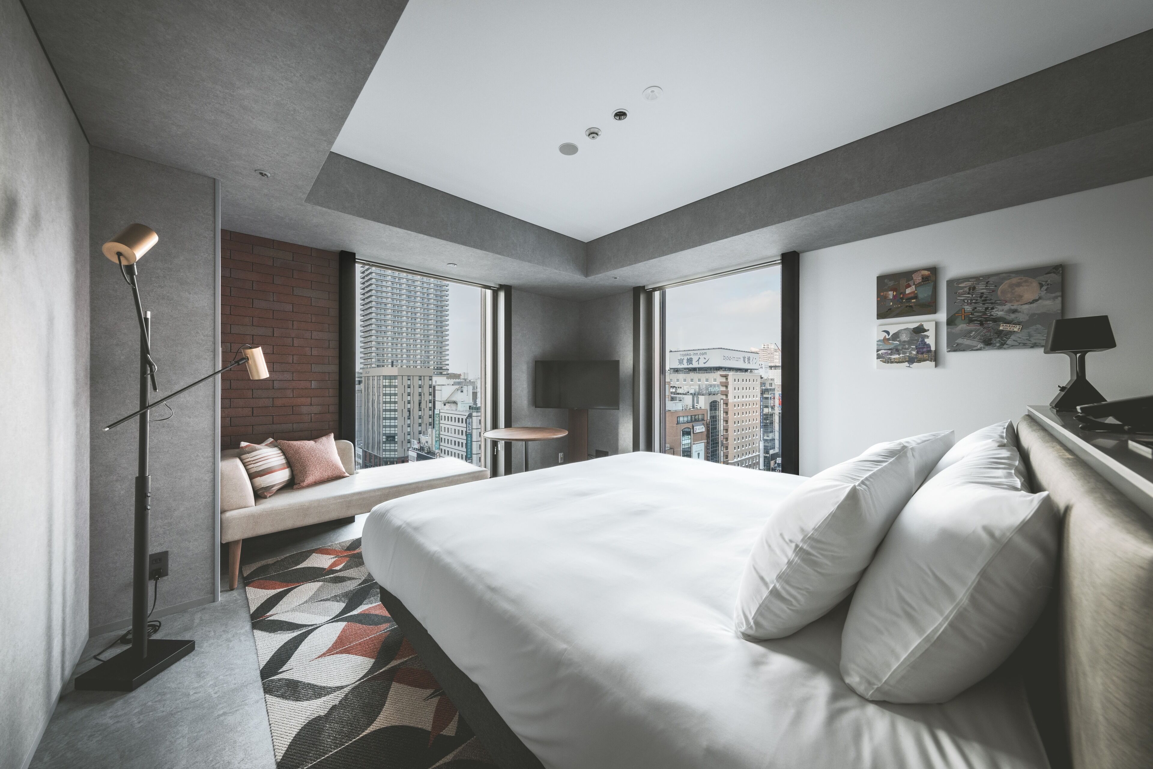 A modern hotel room with a large bed, a seating area with a view, and a cityscape visible through the windows.