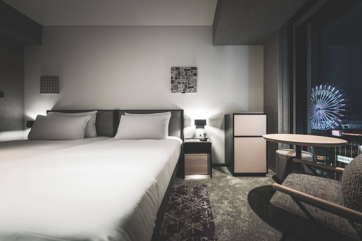 A modern hotel room with a large bed, bedside table, lamp, and a view of a cityscape with a Ferris wheel.