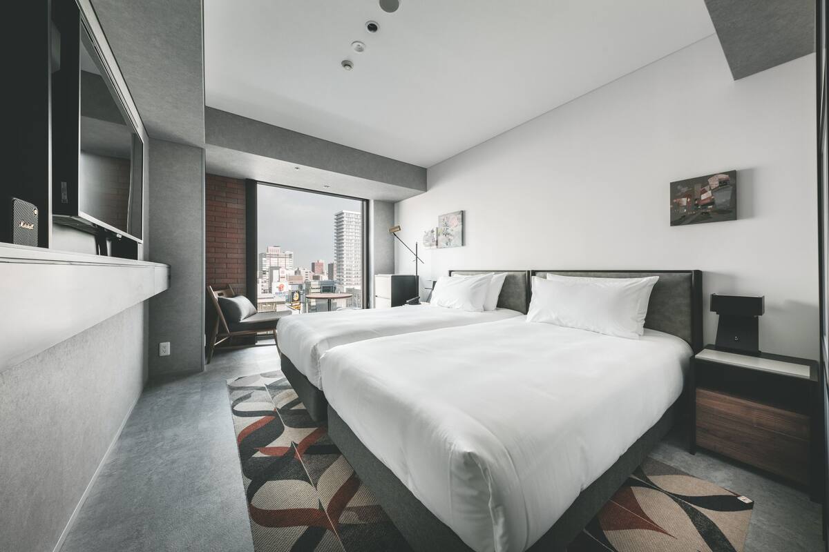 A modern hotel room with two large beds, a city view, a desk, and a large window.