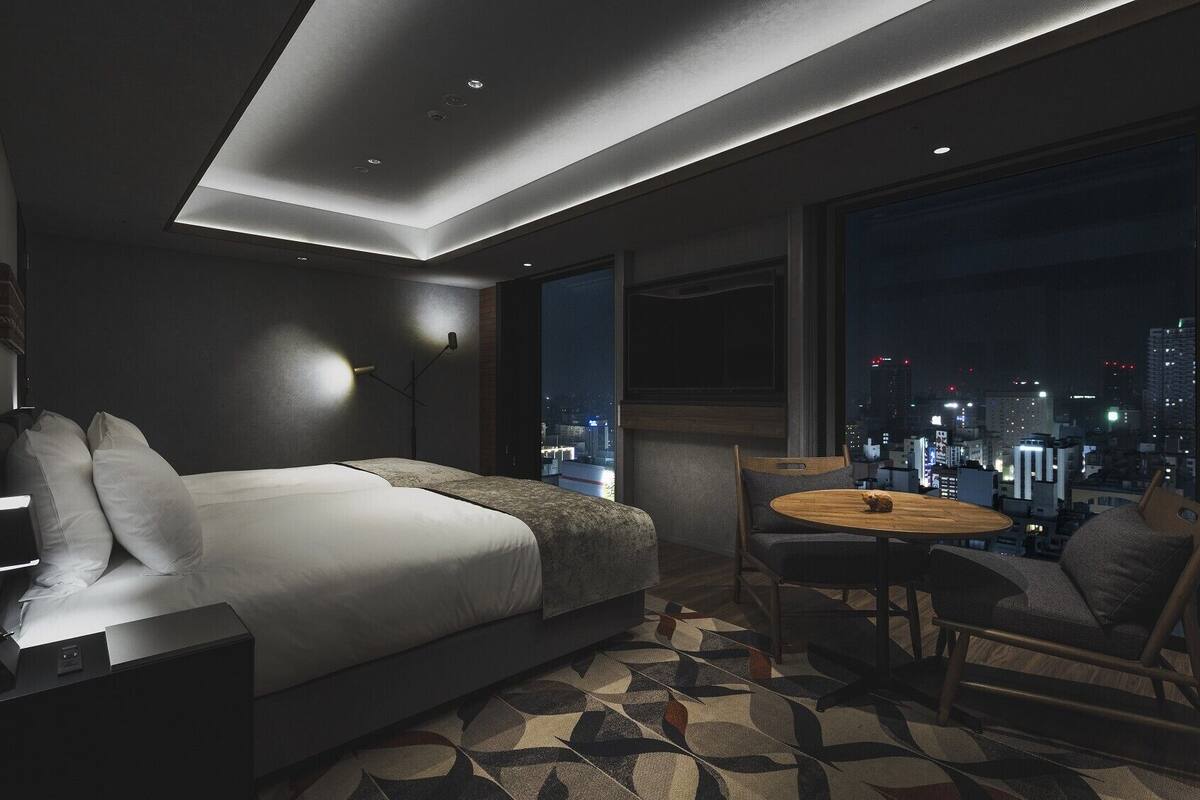 A modern hotel room with a bed, bedside table, lamp, and a city view through large windows.