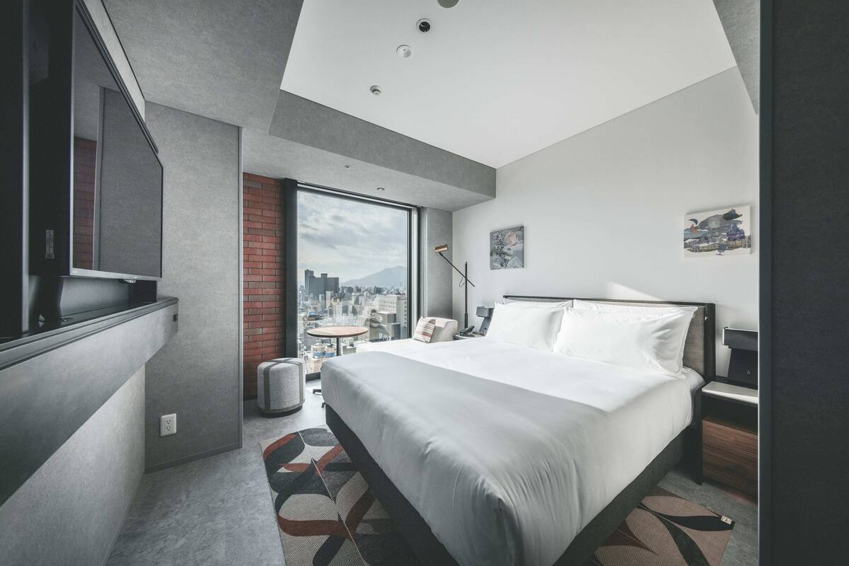 A modern hotel room with a large bed, a city view, a small table, and a chair.