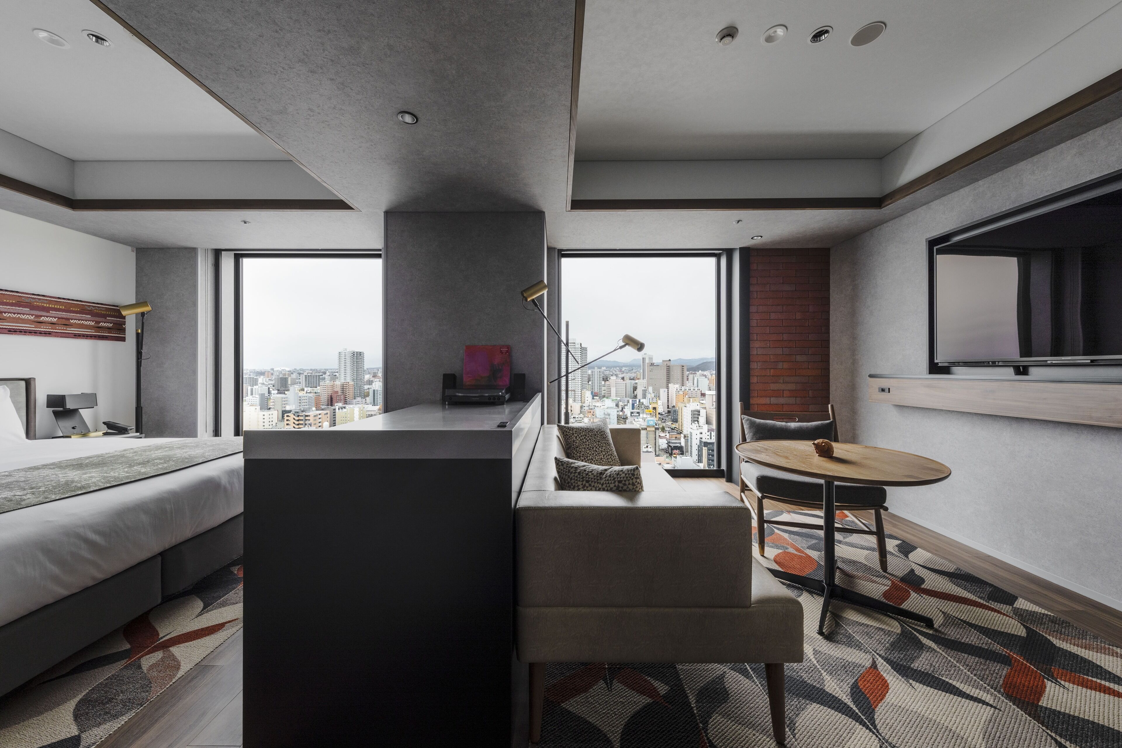 Habitación de hotel moderna con un gran ventanal que ofrece vistas a la ciudad, una zona de estar acogedora y un televisor de pantalla plana montado en la pared.