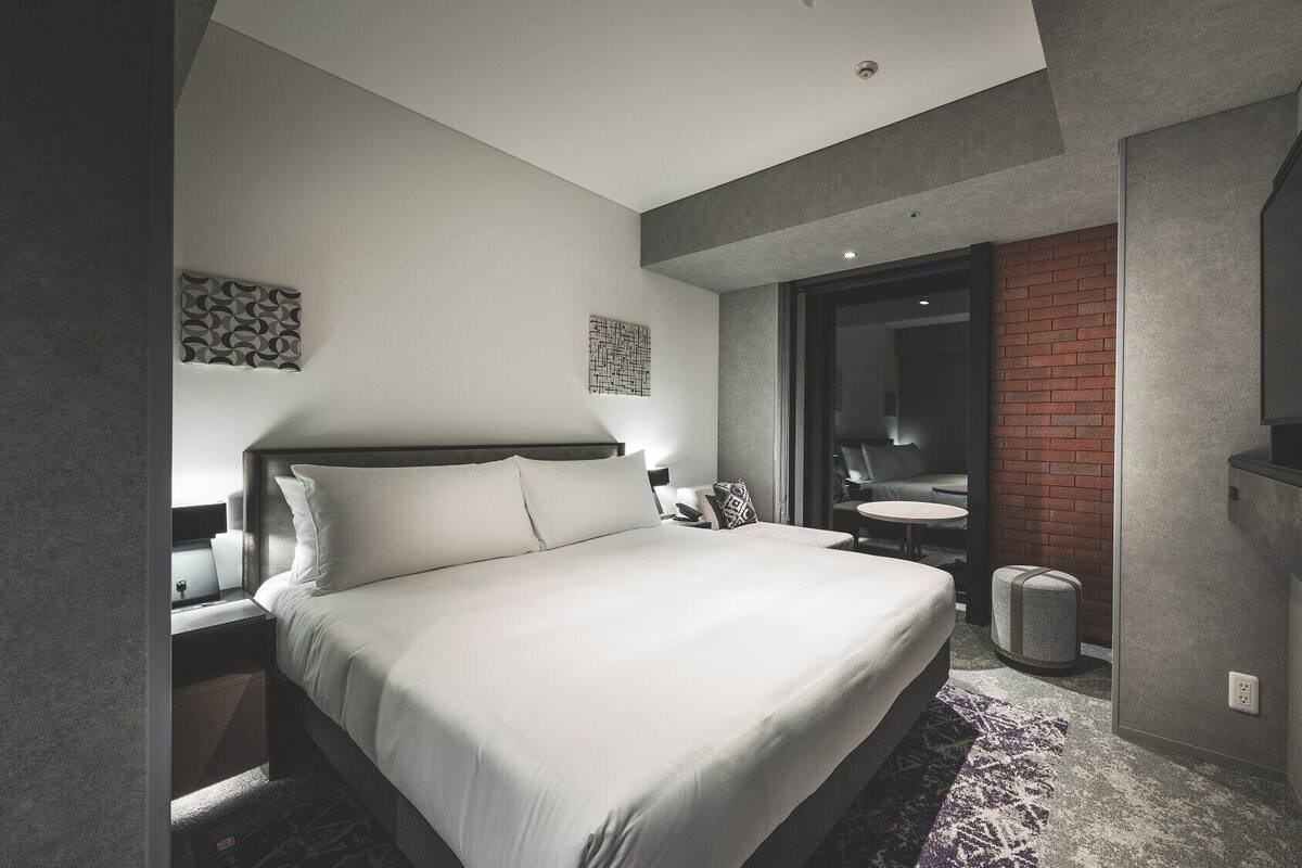 A modern hotel room with a large bed, a desk, a chair, a small round table, and a TV.