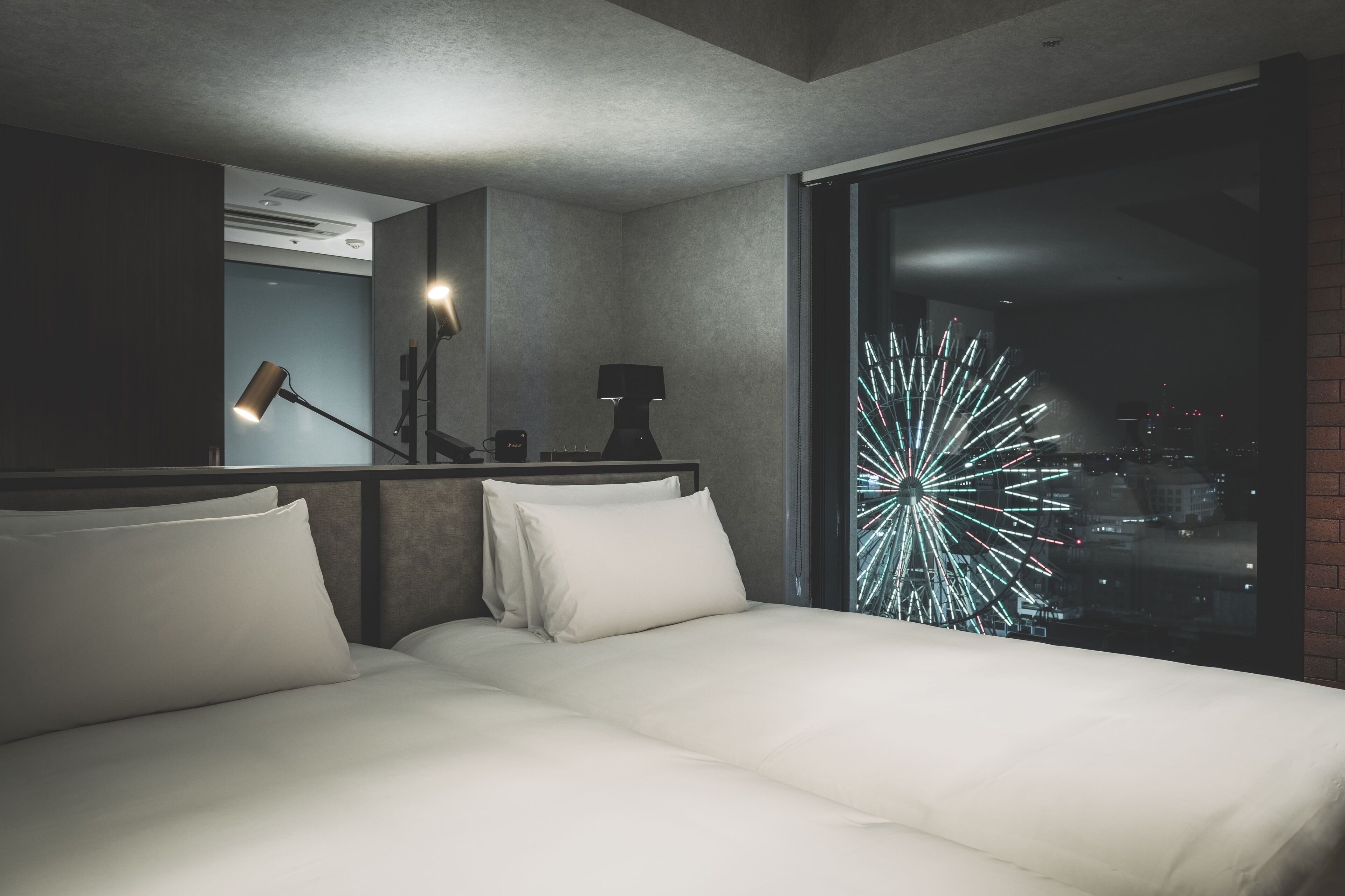 A modern hotel room with a large bed, a nightstand, a lamp, and a view of a cityscape with fireworks.