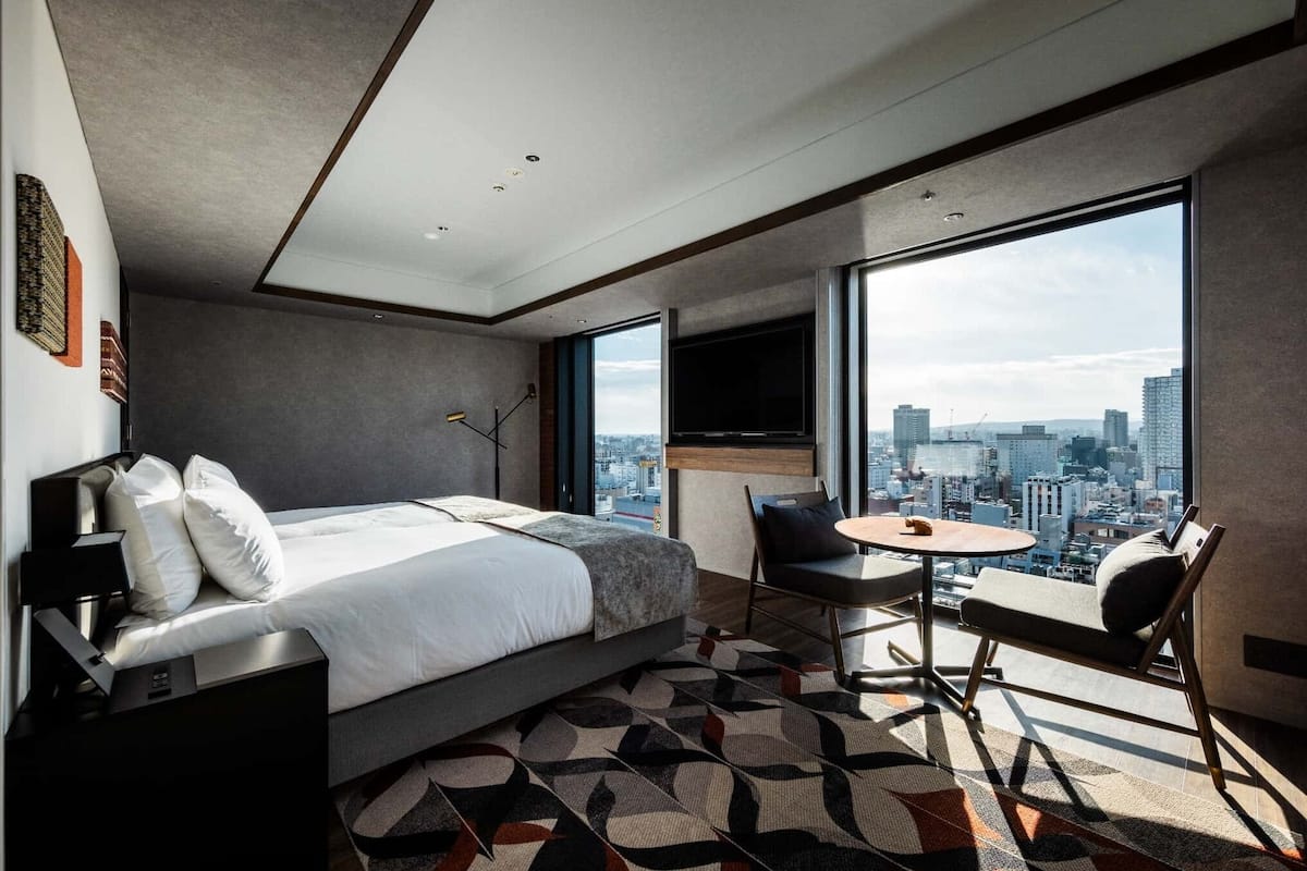A modern hotel room with a large bed, a desk, a chair, and a TV. The room has a city view through large windows.