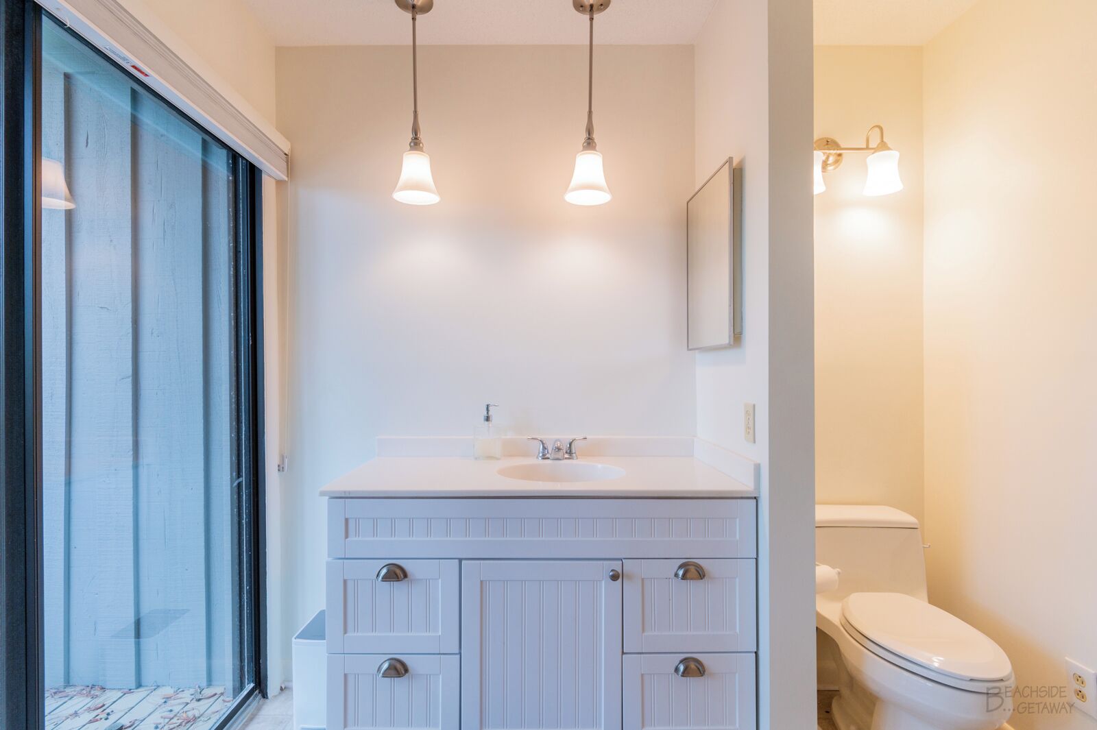 Une salle de bain avec un meuble-lavabo blanc, un évier et des toilettes. Deux lampes suspendues sont fixées au-dessus du meuble-lavabo.