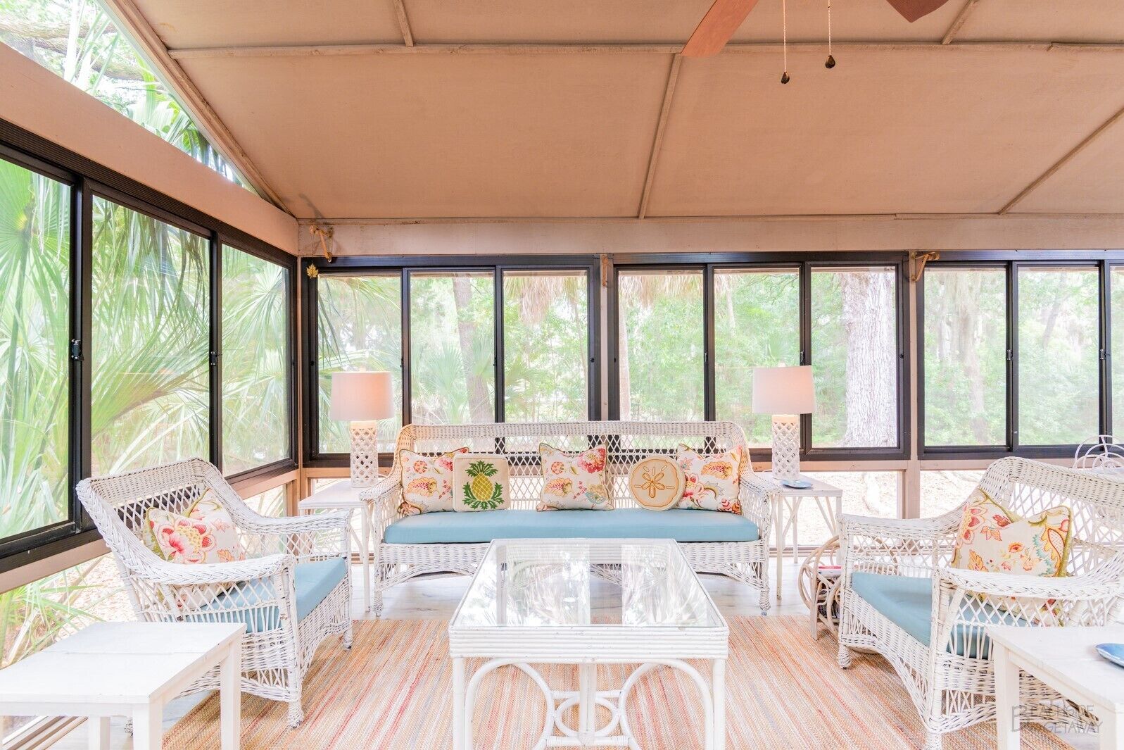 A sunlit room with a white wicker sofa, matching chairs, and a glass coffee table. Large windows offer views of greenery outside.