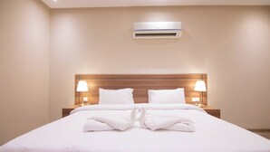 A neatly made bed with white linens and towels arranged in a heart shape, a wooden headboard, bedside lamps, and an air conditioning unit on the wall. - Towlan Jaber Serviced Apartments Jaber Branch (Riyadh)