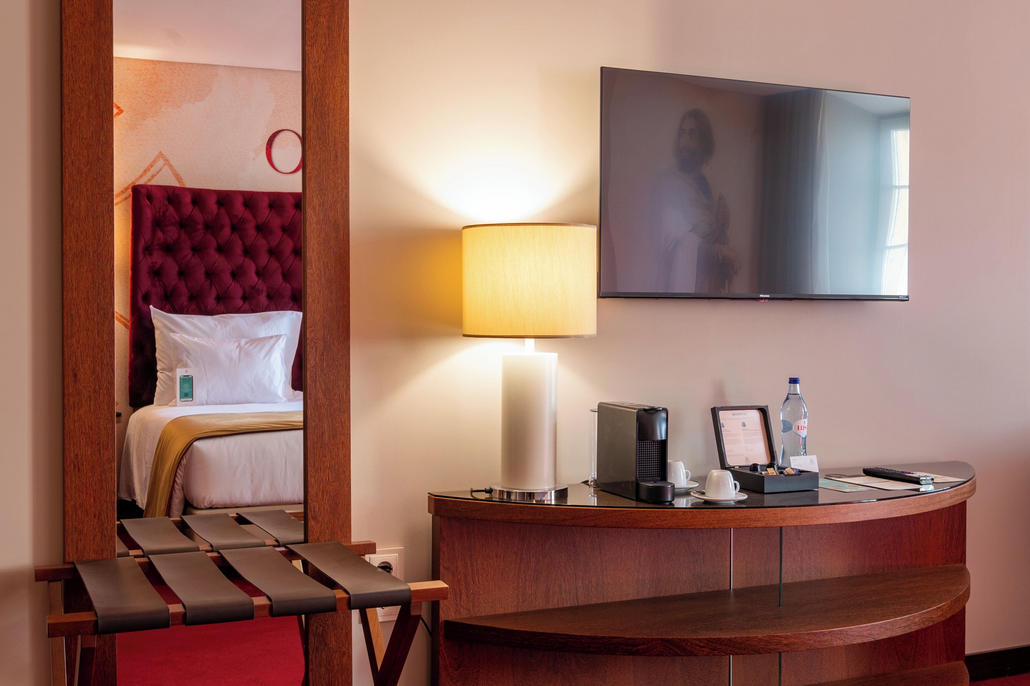 Quarto de hotel com cama, escrivaninha com luminária, televisão e uma cadeira vermelha.