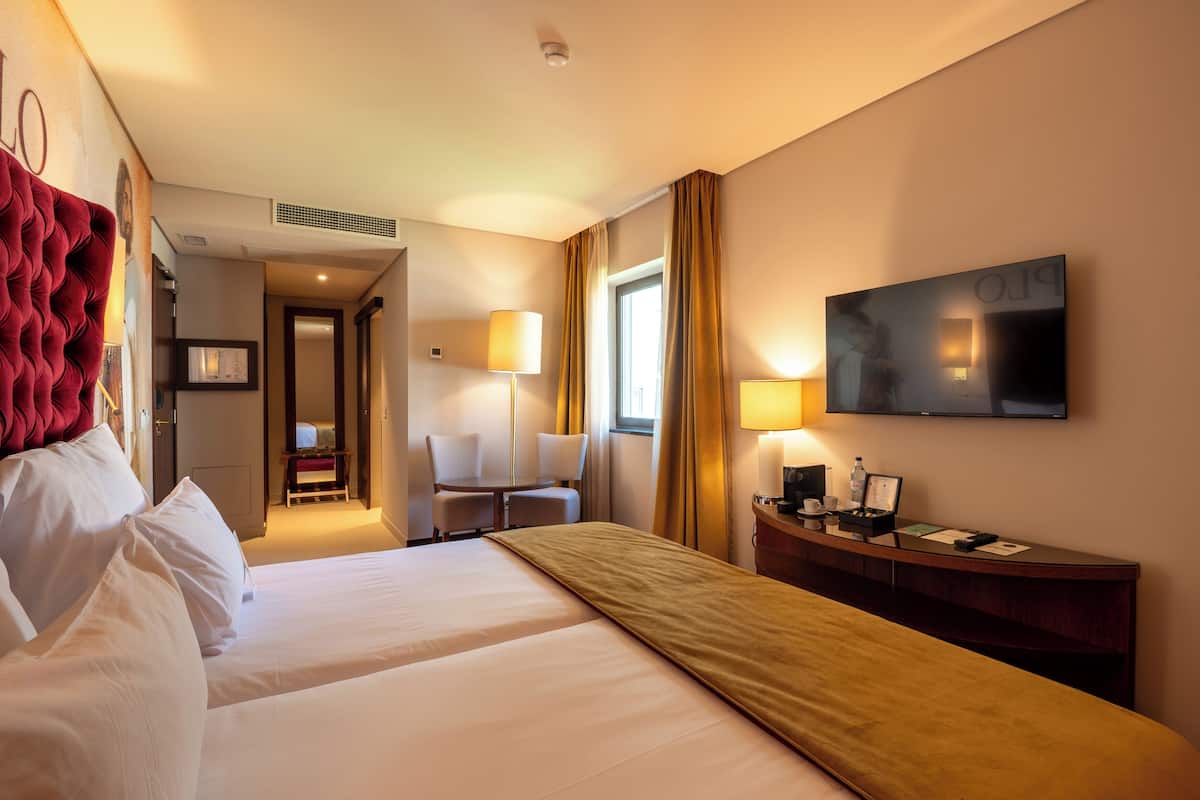 Quarto de hotel com duas camas, uma escrivaninha, uma cadeira, uma televisão e uma janela com cortinas.