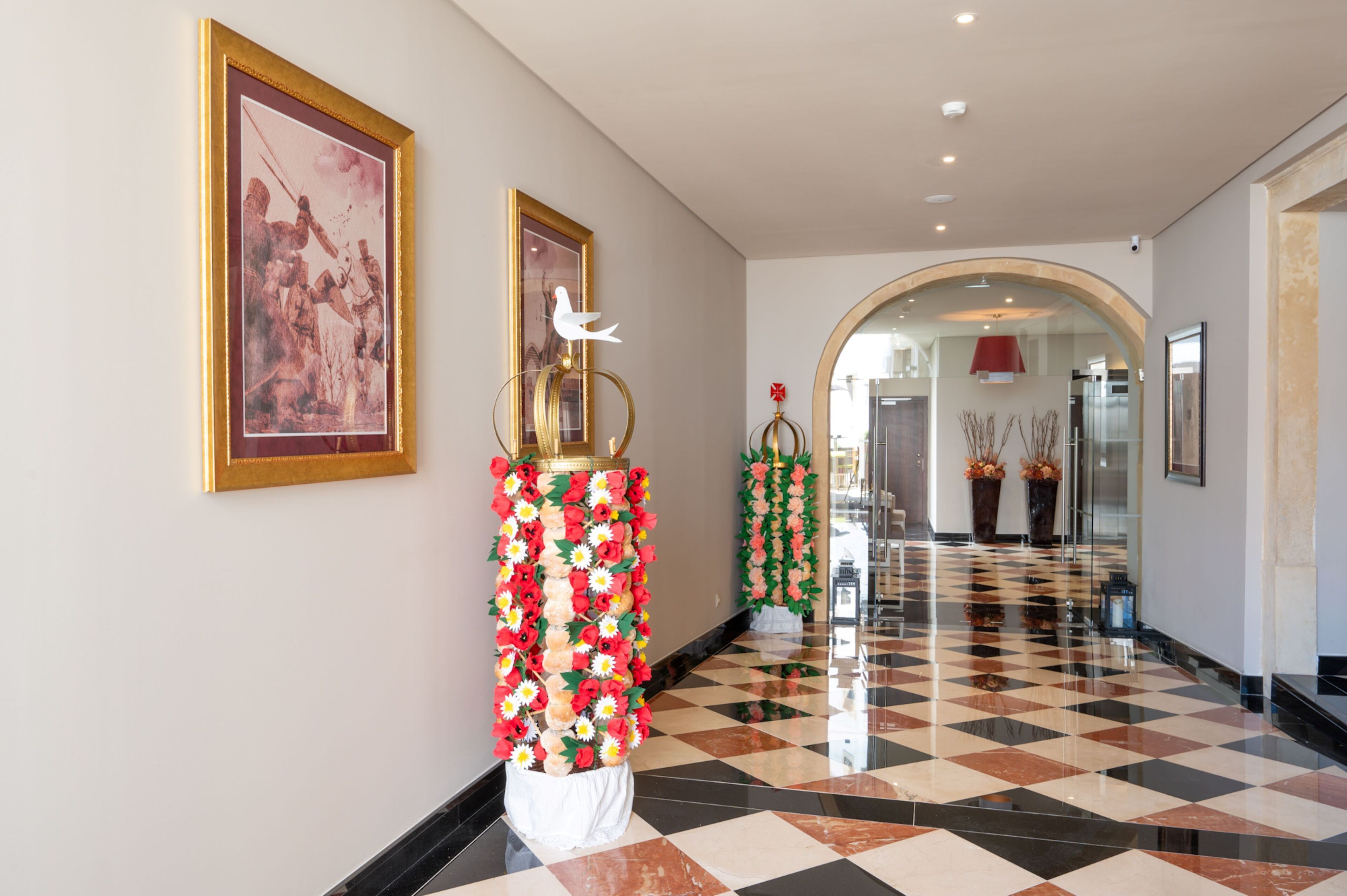 A hotel corridor with a checkered floor, framed artwork on the walls, and decorative floral arrangements.