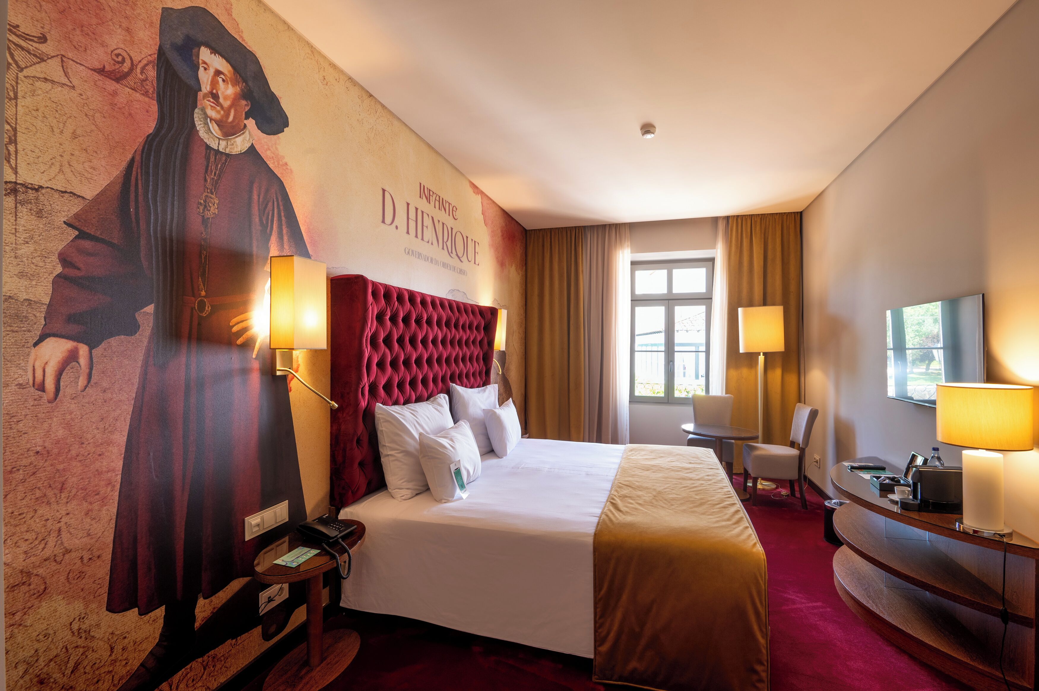 A hotel room with a large portrait on the wall, a tufted headboard, a bed with white linens, a desk with a lamp, and a window with curtains.