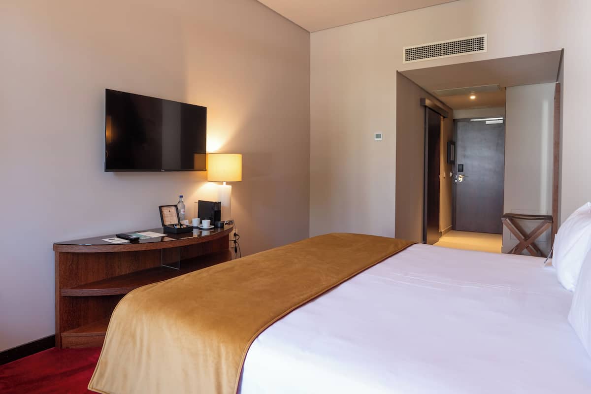 Quarto de hotel com uma cama, uma escrivaninha com luminária, uma TV de tela plana e uma porta que dá para outro quarto.