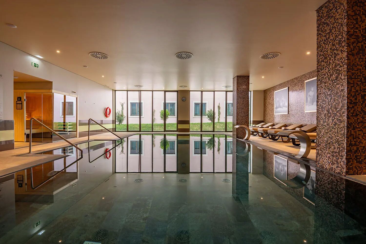 A modern indoor swimming pool with clear glass walls, a sun terrace, and lounge chairs.